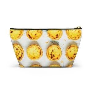May include: A zippered pouch featuring a repeating pattern of golden-brown egg tarts on a white marble background. The accessory has a black top edge and a gold-toned metal zipper pull, showcasing a clear, high-resolution print of the pastries.
