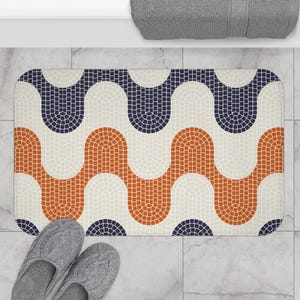 Op de afbeelding: Een rechthoekige badmat met een mozaïekpatroon in marineblauw, oranje en gebroken wit. Het ontwerp heeft een herhalend golfachtig patroon. Grijze pantoffels liggen onderaan de mat.