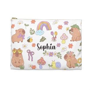 Peut inclure: Une pochette zippée blanche avec un motif répétitif de capybaras bruns, arcs-en-ciel, champignons, escargots, abeilles, fleurs et nœuds violets. Le prénom Sophia est imprimé en écriture noire au centre de la pochette parmi les illustrations.
