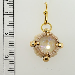 Pode incluir: Um único brinco de contas com um gancho dourado. O brinco apresenta um cristal iridescente central rodeado por contas bege e detalhes dourados. O brinco mede aproximadamente 2 cm de comprimento.