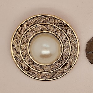 May include: A gold-toned, circular brooch with a large, pearlescent center. The brooch features a detailed, textured design around the pearl. A US penny is next to the brooch for size comparison.