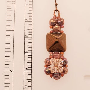 May include: A handcrafted earring featuring a copper-colored square bead, surrounded by smaller beads in shades of copper and pink. The earring is approximately 3 inches long, with a decorative floral-shaped cluster at the bottom.