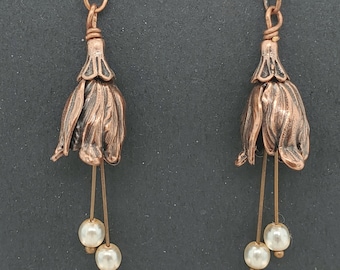 Pode incluir: Um par de brincos cor de cobre. Cada brinco apresenta um design de flor em forma de sino com duas mechas penduradas, cada uma segurando uma pérola branca. Os brincos têm um fecho de gancho.