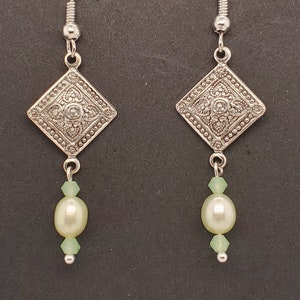 May include: Silver dangle earrings with a square-shaped pendant featuring an intricate design. The pendant is attached to a single pearl bead and two green crystal beads.