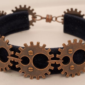 Op de afbeelding: Een steampunk-armband met bronskleurige tandwielen die met elkaar verbonden zijn. De tandwielen zijn bevestigd aan een donkerblauwe fluwelen band. De armband heeft een sluiting.