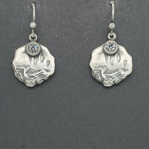 May include: A pair of silver dangle earrings with a textured, irregular shape and a single clear rhinestone in the centre of each earring.