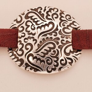 Puede incluir: Un colgante circular plateado con un patrón ornamental y ondulado es el punto focal. Está conectado a una estrecha correa de cuero marrón rojizo, formando una pulsera o accesorio similar. El diseño es detallado y llamativo, ideal para cualquier ocasión.