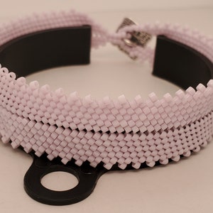 Op de afbeelding: Een lichtpaarse kralenarmband met een zwarte binnenkant. De armband is gemaakt van kleine, vierkante kralen die aan elkaar zijn geweven. De sluiting is zilverkleurig.