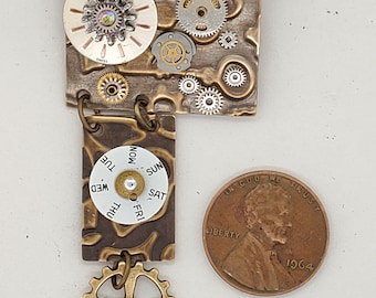 May include: A steampunk-style jewelry piece with a clock face, gears, and a day of the week dial. The piece is made of metal and has a brown and gold finish. A penny is included for scale.