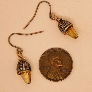 May include: A pair of gold-toned earrings with a textured, acorn-shaped design and a faceted, amber-colored crystal. The earrings are displayed next to a U.S. penny from 1964.