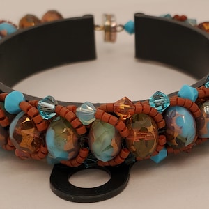 May include: A beaded bracelet with a black base, adorned with turquoise and bronze beads. The bracelet, with a magnetic clasp, showcases a variety of bead sizes and shapes, creating a textured and colourful accessory.