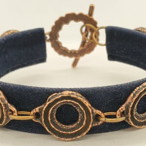 May include: A dark blue bracelet with copper-colored circular links. The links are connected by small gold-colored rings. The bracelet has a toggle clasp. The band of the bracelet is a dark blue fabric.