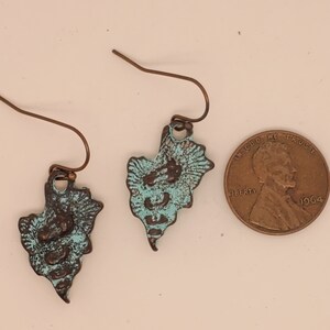 May include: Handmade earrings with a distinctive, textured design. The earrings have a teal and bronze colour scheme, with a weathered appearance. The earrings are shown next to a penny for size comparison.