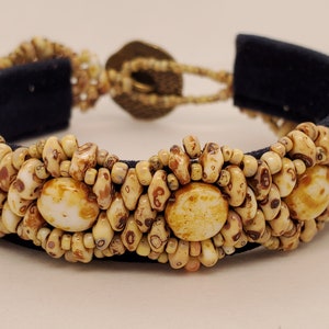 May include: A beaded bracelet featuring a dark blue fabric base, adorned with cream and brown-toned beads. The bracelet has a central design of larger, round beads and a clasp closure. The overall design is intricate and detailed.
