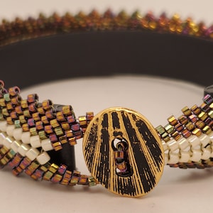 Puede incluir: Una pulsera de cuentas con base negra y cierre de botón dorado. La pulsera presenta un patrón de cuentas blancas, doradas e iridiscentes. El cierre de botón es dorado con un diseño negro texturizado.