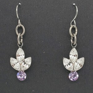 May include: A pair of silver-toned dangle earrings. Each earring features a leaf-shaped design with three clear crystals and a round, light purple crystal at the bottom. The earrings hang from a simple hook.