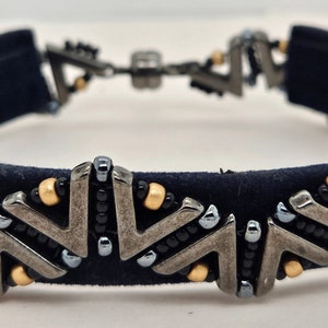 May include: A navy blue velvet bracelet with a geometric pattern of silver and gold beads. The bracelet is adorned with small silver triangles and gold beads.