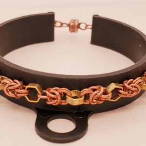 Könnte beinhalten: Ein Armband aus Kupfer und Messing mit einem gewebten Design und sechseckigen Akzenten. Das Armband wird auf einem schwarzen Ständer präsentiert.