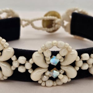 Könnte beinhalten: Ein Perlenarmband mit einem schwarzen Band, cremefarbenen Perlen und schillernden Kristallen. Das Armband hat ein zentrales Blumenmuster und einen Knopfverschluss. Die Perlen sind in einem dekorativen Muster angeordnet.