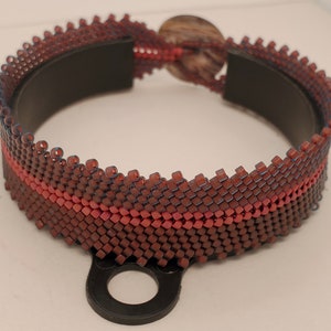 Może przedstawiać: Bransoletka z brązowych i czerwonych koralików z czarną metalową klamrą. Bransoletka ma jeden czerwony pasek przechodzący przez brązowe koraliki.