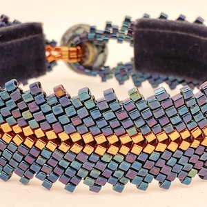 Könnte beinhalten: Ein breites Perlenarmband mit dunkelblauem Samtrücken. Das Armband hat ein Zickzackmuster aus irisierenden blauen und goldenen Perlen. Der Verschluss ist eine runde, marmorierte Perle mit kupferfarbenen Metallakzenten.