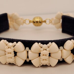 May include: Cream-colored beaded bracelet with a dark blue velvet backing. The bracelet features a repeating pattern of cream-colored beads and a gold-colored magnetic clasp. A stylish piece of jewelry.