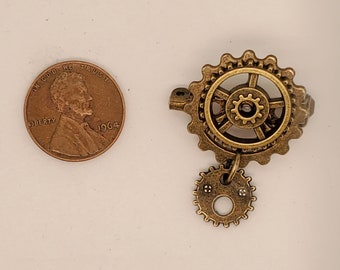 May include: A bronze-colored steampunk-style ring with a gear design. The ring features a large gear with a smaller gear hanging below. A U.S. penny from 1964 is next to the ring for size comparison.
