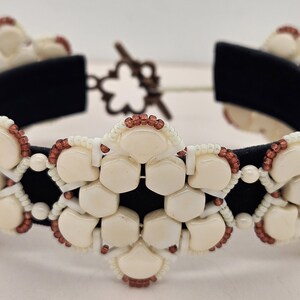 Puede incluir: Una pulsera de cuentas color crema con un respaldo de terciopelo negro. La pulsera presenta un diseño floral con pequeñas cuentas marrones y blancas. El cierre es un broche de palanca en forma de flor, marrón oscuro.