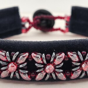 Op de afbeelding: Een zwarte fluwelen armband met een herhalend bloemmotief van metallic zilveren en doorschijnende rode kralen. De armband wordt vastgezet met een donkere ronde knoopsluiting die verbonden is door een dunne rode koordlus aan de achterkant.