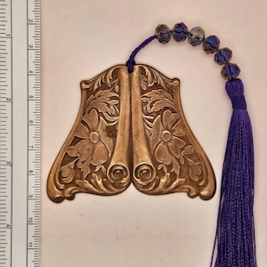 May include: A gold-colored bookmark with an ornate design featuring scrolls and flowers. It has a purple tassel and a string of iridescent purple beads.