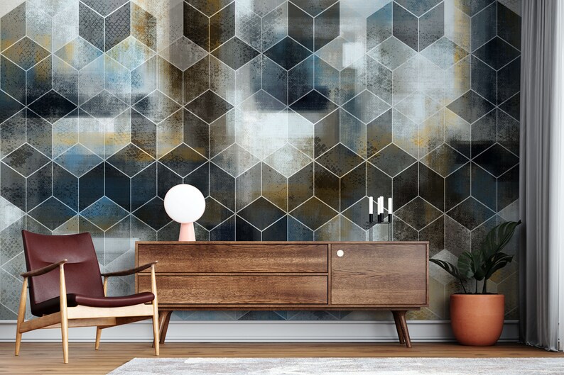 May include: A brown wooden media console with two drawers and a single door. The console is in front of a wall with a geometric pattern of blue, gray, and gold cubes. A white lamp sits on the console, and a potted plant is on the right side of the console.