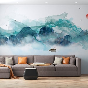 May include: A watercolor painting of a mountain landscape with a boat in the foreground. The mountains are painted in shades of blue and green, and the sky is a light blue. The painting is on a white wall in a living room with a gray sectional sofa and a coffee table.