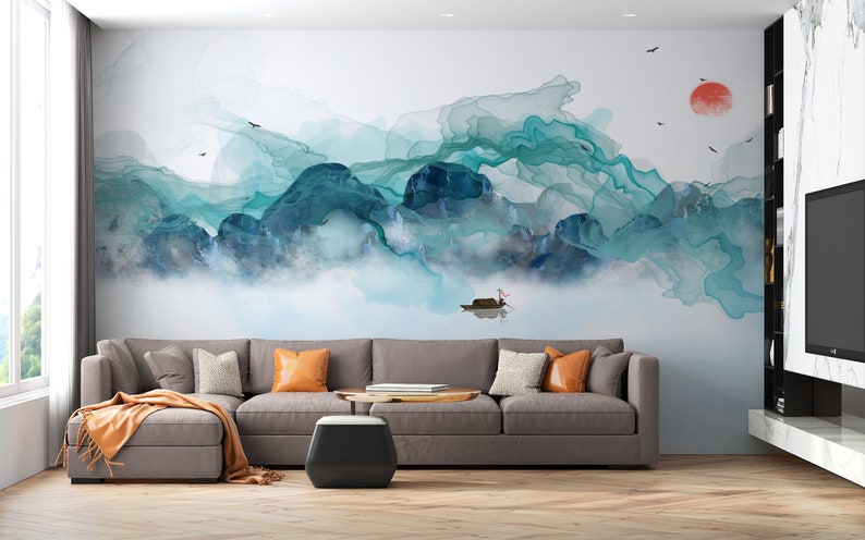 May include: A watercolor painting of a mountain landscape with a boat in the foreground. The mountains are painted in shades of blue and green, and the sky is a light blue. The painting is on a white wall in a living room with a gray sectional sofa and a coffee table.