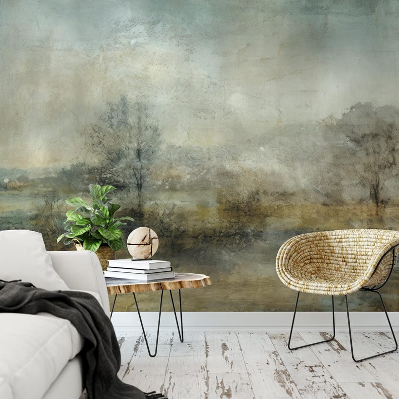May include: A muted green and brown abstract landscape painting with a hazy, atmospheric feel. The painting is displayed on a wall in a living room setting with a white couch, a wooden side table, and a wicker chair.