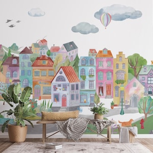 Puede incluir: Un mural de acuarela que representa una ciudad colorida con casas, árboles y un globo aerostático. El mural está pintado en una pared blanca y presenta una variedad de colores, incluyendo rosa, azul, amarillo, verde y rojo. El mural es una adición caprichosa y encantadora a cualquier habitación.