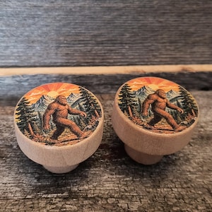 May include: Two round wooden cabinet knobs featuring a printed graphic of a brown Bigfoot creature walking through a forest with snow-capped mountains and a bright orange sun in the background. The knobs have a natural light wood finish.