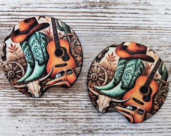 May include: Two circular car coasters featuring a western-themed illustration. The design includes a brown cowboy hat, turquoise patterned cowboy boots, an acoustic guitar, a steer skull, a wooden wagon wheel, and an orange vintage pickup truck.