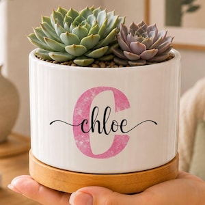 May include: A white cylindrical ceramic planter with vertical ridges, featuring a large pink watercolour letter C and the name chloe in black script. The planter sits on a circular wooden base and contains two green and purple succulent plants.