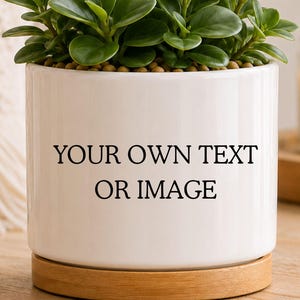 May include: A white cylindrical ceramic plant pot with a light wood base, featuring the black printed text "YOUR OWN TEXT OR IMAGE" on the front. The pot contains a green leafy plant with rounded, succulent-like foliage and brown soil.