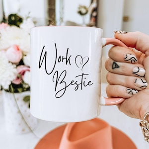 May include: White ceramic mug with the words "Work Bestie" in black script. The mug has a pink interior and handle. The mug is held in a hand with decorated nails, and is next to a vase of flowers.