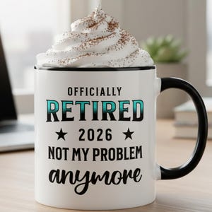 Op de afbeelding: Een witte keramische mok met een zwart handvat en rand, gevuld met slagroom en cacaopoeder. De mok heeft de tekst "OFFICIALLY RETIRED 2026 NOT MY PROBLEM anymore" in zwarte en blauwgroene letters.