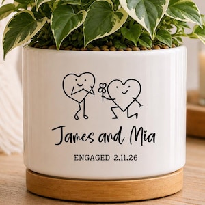 May include: A white cylindrical ceramic planter with a wooden base containing a variegated green and white heart-leaf plant. The front features a black line drawing of two heart characters, the text "James and Mia", and "ENGAGED 2.11.26".