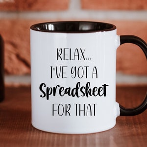 Op de afbeelding: Witte keramische koffiemok met een zwarte rand en handvat. De mok heeft zwarte tekst met de tekst "Relax... I've got a Spreadsheet for that."