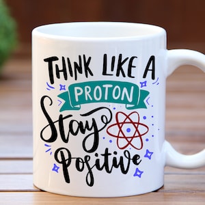 May include: White ceramic mug with a black and teal design that says "Think like a proton stay positive". The design also includes a red and blue atom symbol.