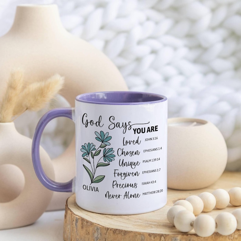 May include: White ceramic mug with a purple interior and handle. The mug features the text "God Says You Are" with a floral design and inspirational words like "Loved" and "Chosen". The mug is personalized with the name "Olivia".