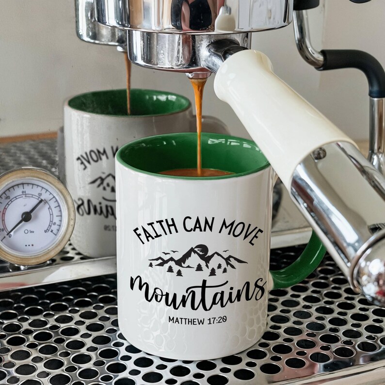 May include: A white ceramic mug with a green interior featuring the black text FAITH CAN MOVE mountains and a mountain landscape graphic with the citation MATTHEW 17:20. Espresso pours from a machine into the mug, which sits on a metal drip tray.