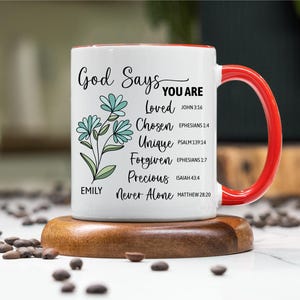 May include: White ceramic mug with a red handle and rim, featuring the text "God Says You Are" and inspirational words. The mug has a floral design and is on a wooden coaster. The mug is approximately 4 inches tall.