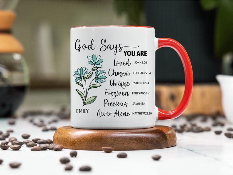 May include: White ceramic mug with a red handle and rim, featuring the text "God Says You Are" and inspirational words. The mug has a floral design and is on a wooden coaster. The mug is approximately 4 inches tall.