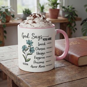 May include: White ceramic mug with a pink handle and rim, filled with a whipped topping and cocoa powder. The mug features the text "God Says You Are" with inspirational words and floral illustrations. A Bible rests on a wooden table.