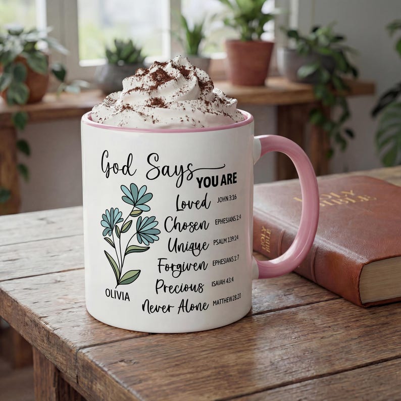 May include: White ceramic mug with a pink handle and rim, filled with a whipped topping and cocoa powder. The mug features the text "God Says You Are" with inspirational words and floral illustrations. A Bible rests on a wooden table.
